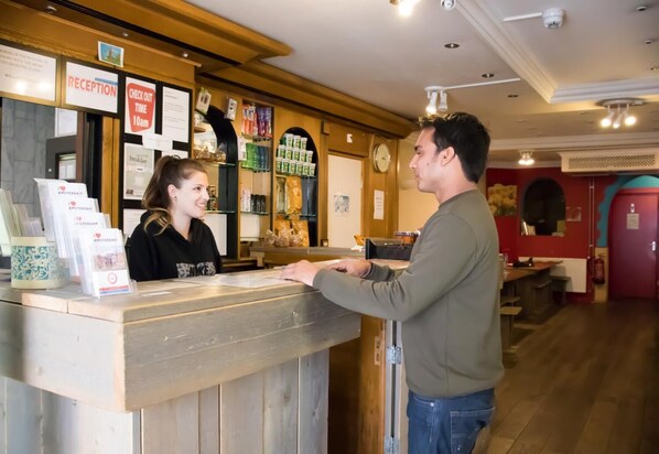 A reception desk with a wooden counter, a clock, and various signs including "RECEPTION" and "CHECK OUT TIME 10am". - Crossroads Hostel Amsterdam (Amsterdam)