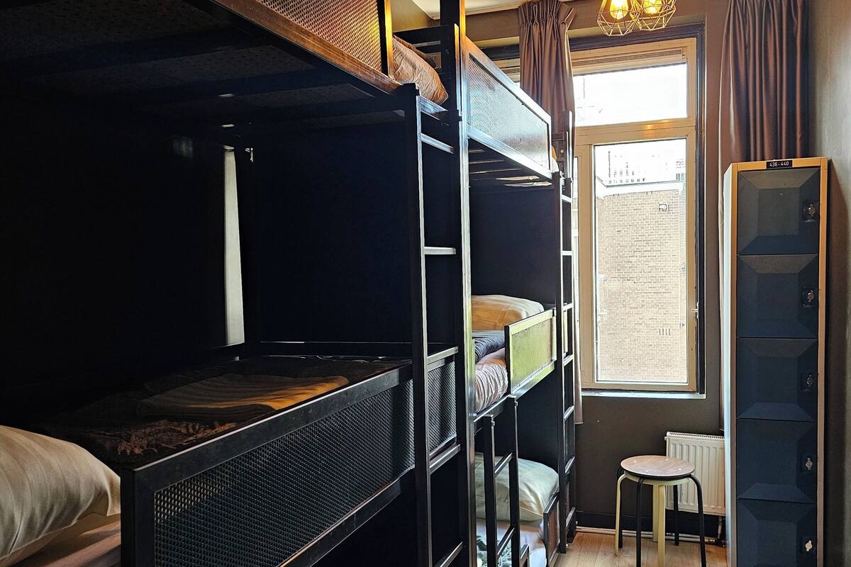 A dormitory room with bunk beds, a small stool, a radiator, and a window with a view of the outside.