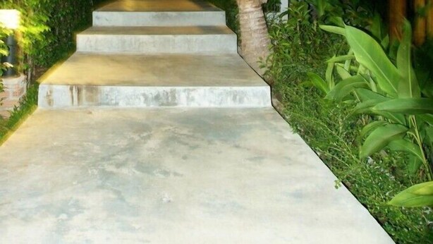 A concrete pathway leading to a wooden building with a thatched roof, surrounded by lush greenery and palm trees. - The Fisherman's Chalet (Koh Samui)