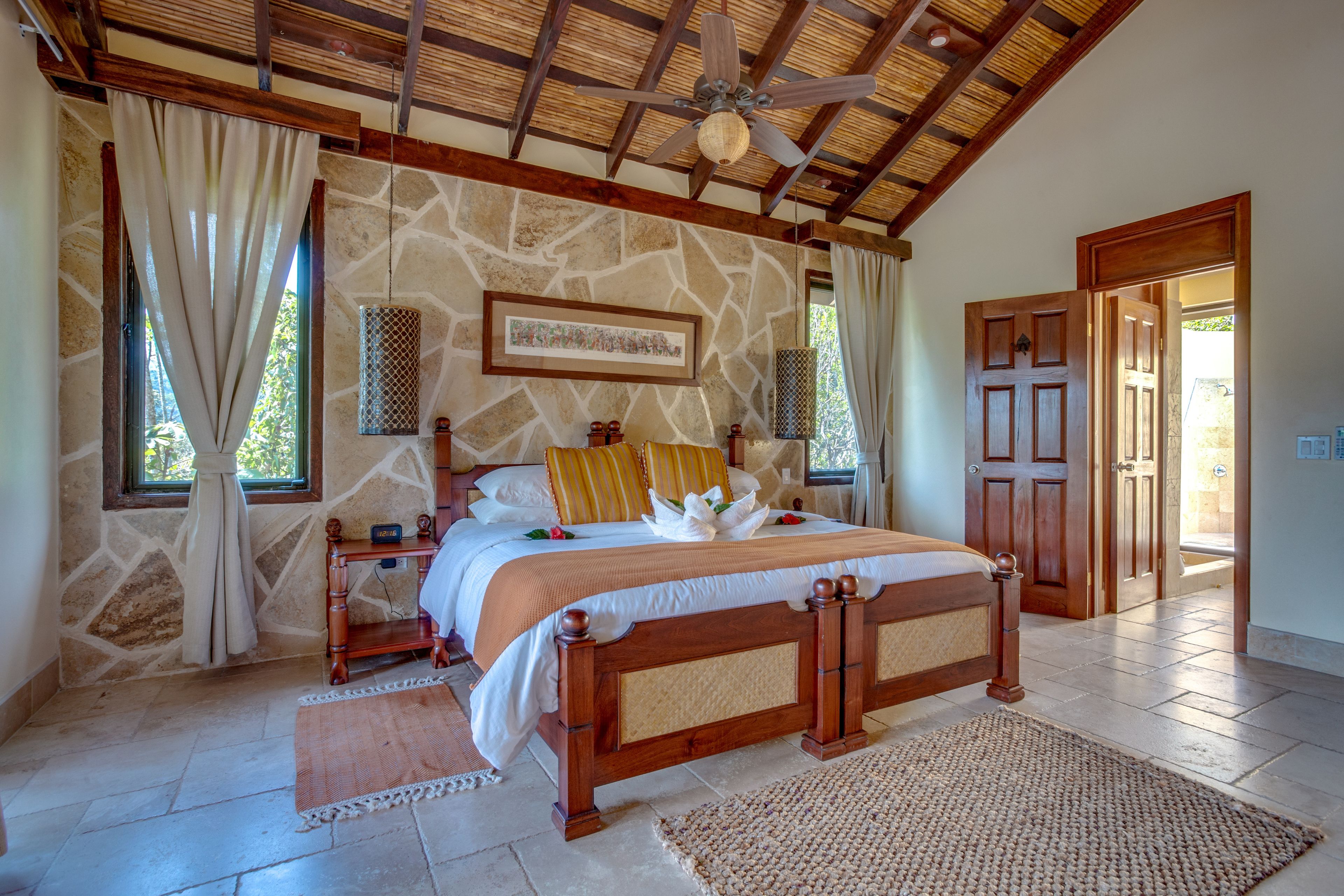 A bedroom with a stone wall, a wooden bed, a ceiling fan, and a large window with curtains.
