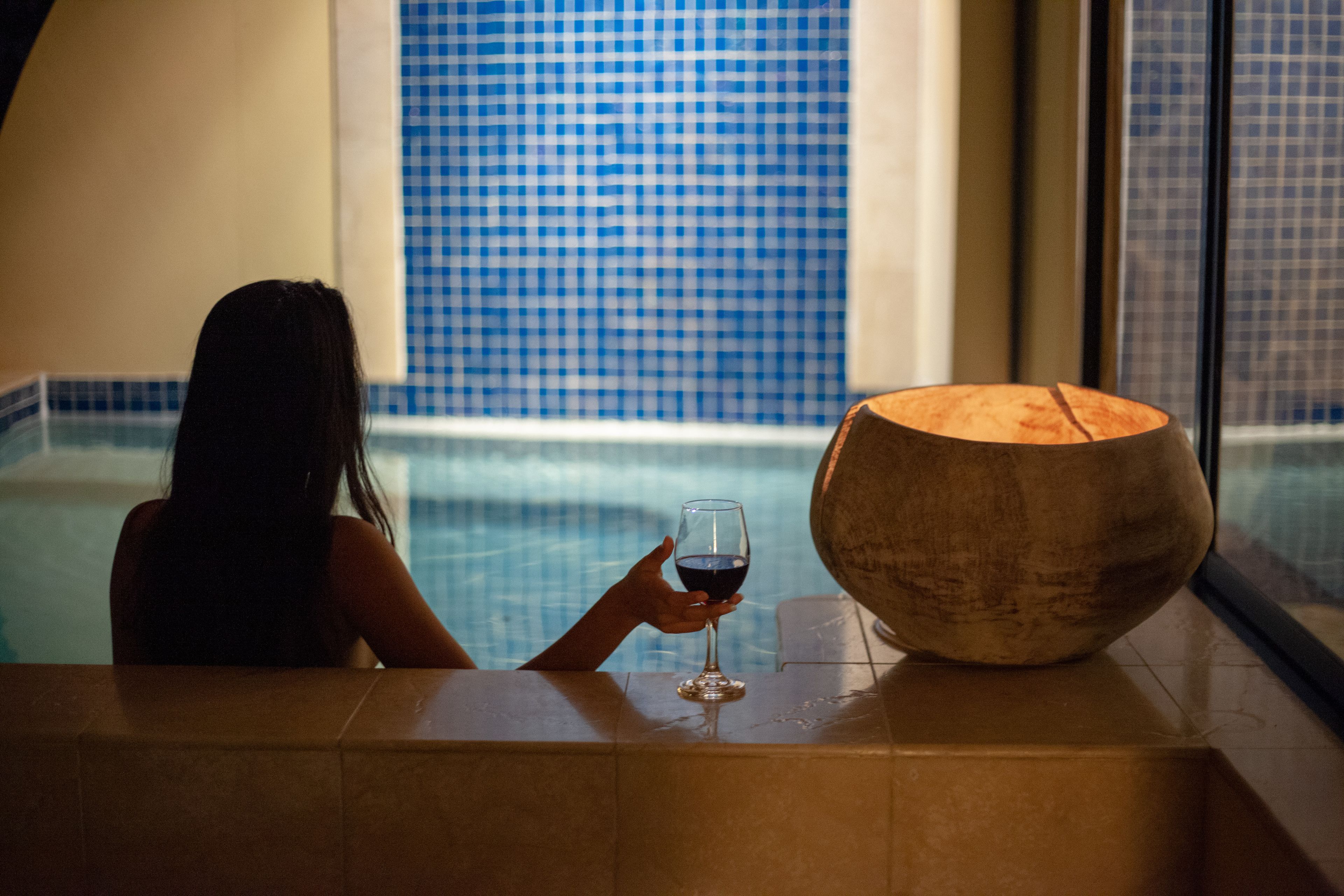 A person sitting by a pool with a glass of red wine, a wooden bowl, and a checkered blue and white tiled wall in the background.