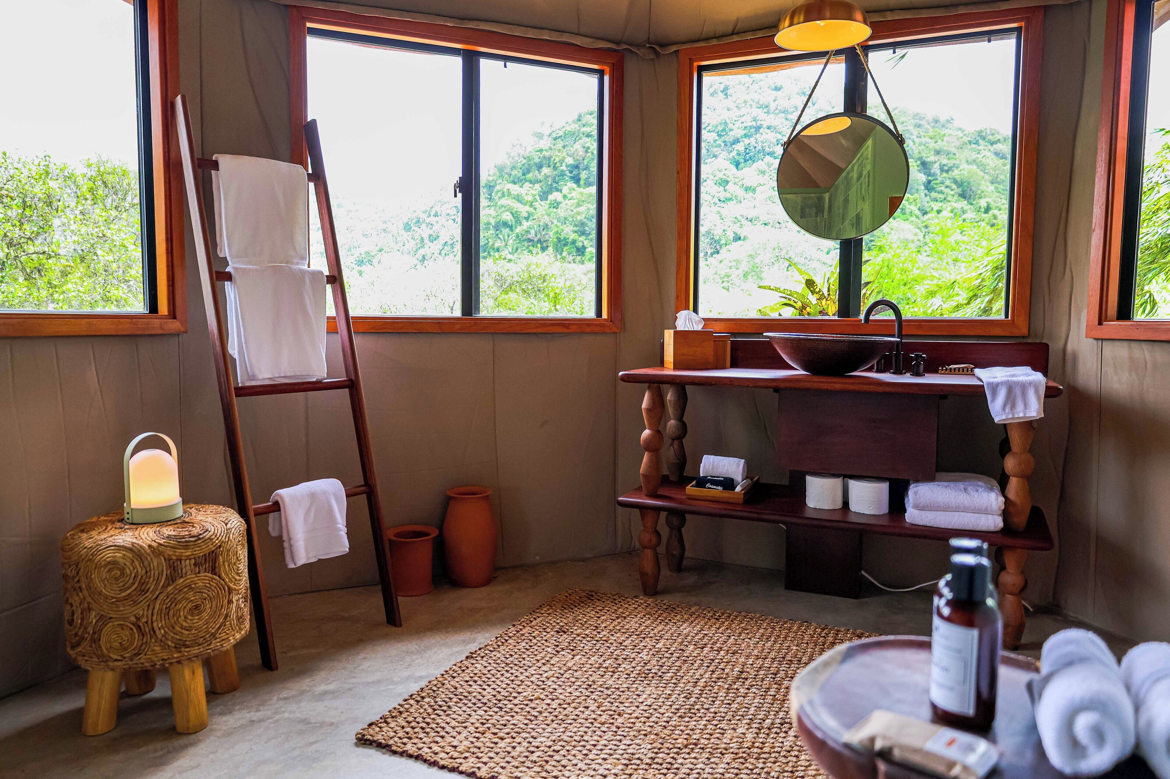 A bathroom with a wooden ladder, a woven stool with a lantern, a sink with a mirror, and towels.