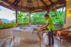 Un cuarto de terapia de masaje con techo de madera, una camilla y vistas a exuberante vegetación y montañas.