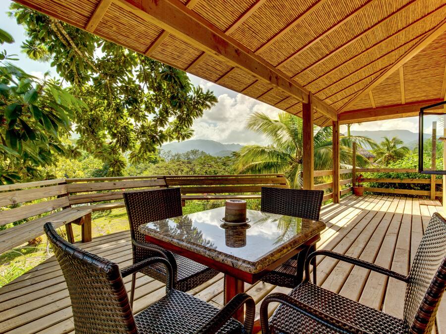 Un espacio de estar al aire libre cubierto, con una mesa y sillas, rodeado de abundante vegetación y vistas a montañas distantes.