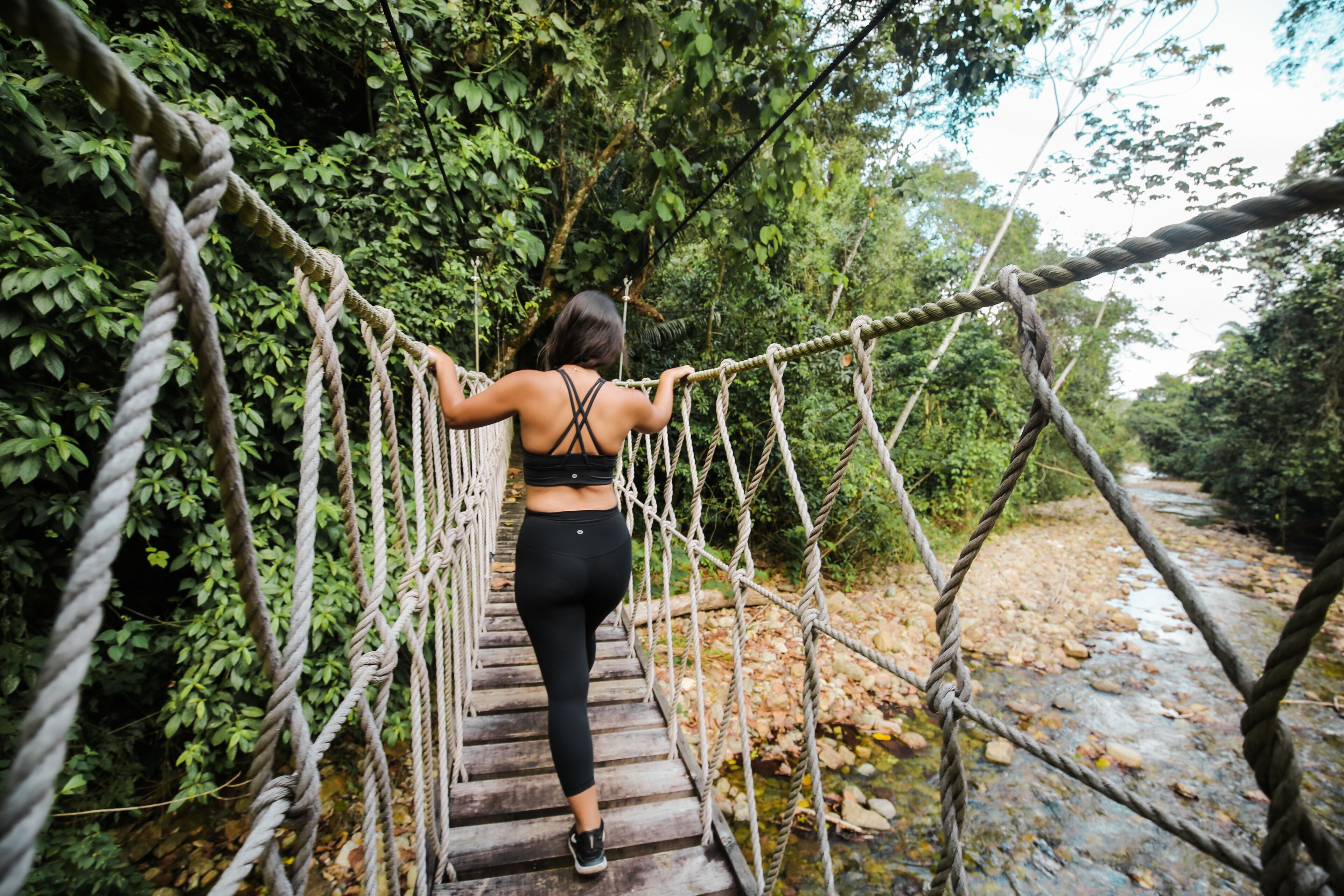 Una persona caminando sobre un puente colgante en un área boscosa con un arroyo debajo.