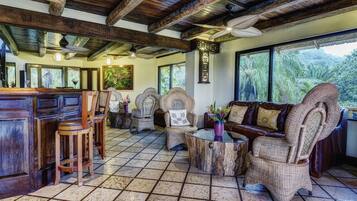 A spacious living area with a high ceiling, wooden beams, and large windows offering a view of lush greenery.