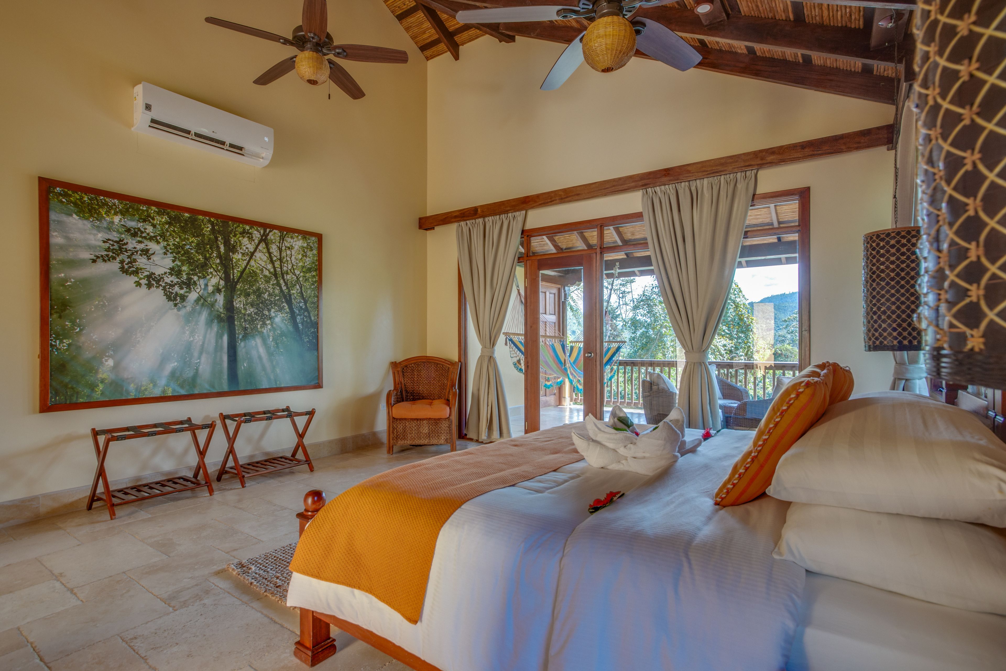 A bedroom with a bed, a ceiling fan, an air conditioning unit, a large painting, a wicker chair, and a view of the outdoors.