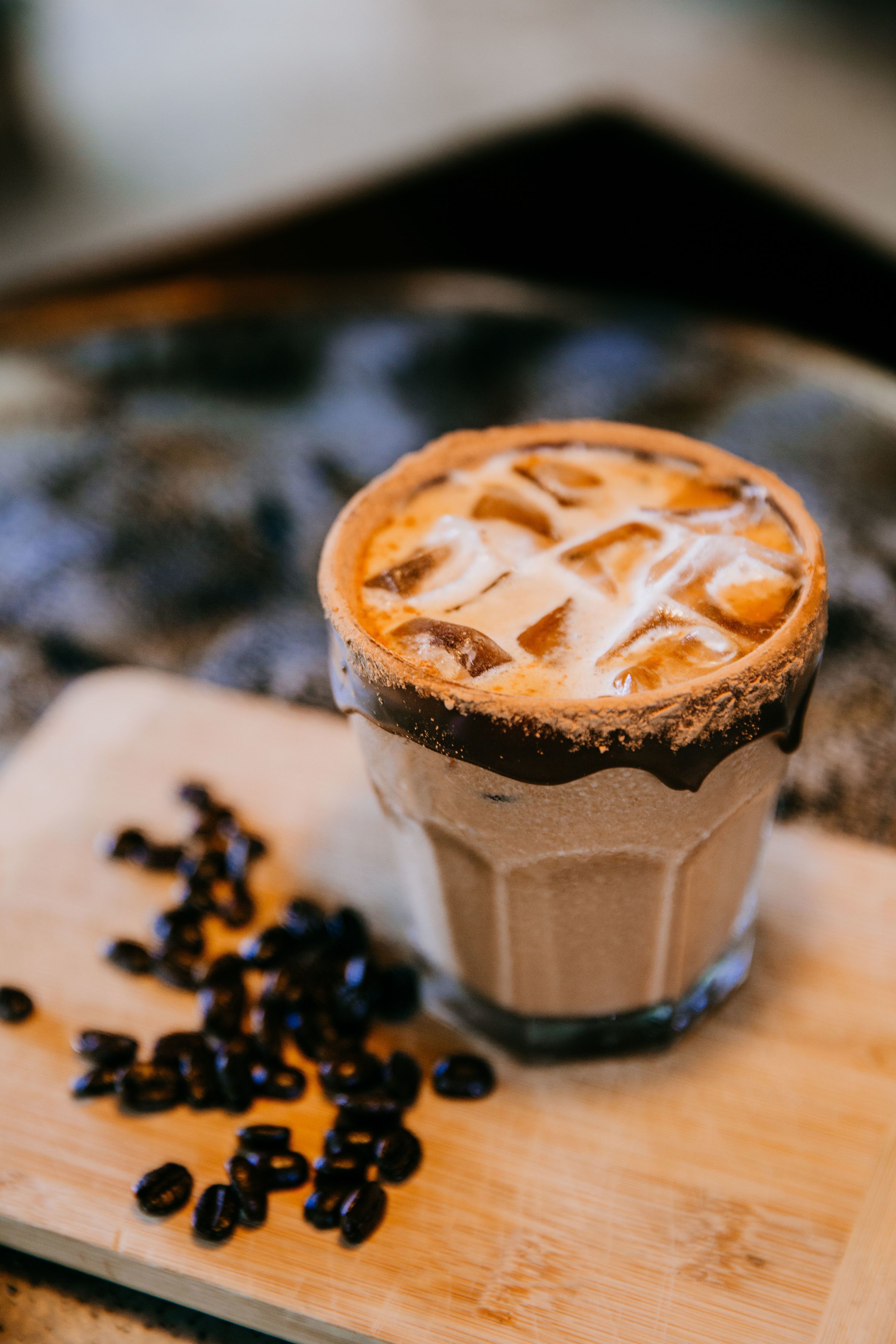 A glass of iced coffee with a caramelized top, placed on a wooden surface with scattered coffee beans beside it.