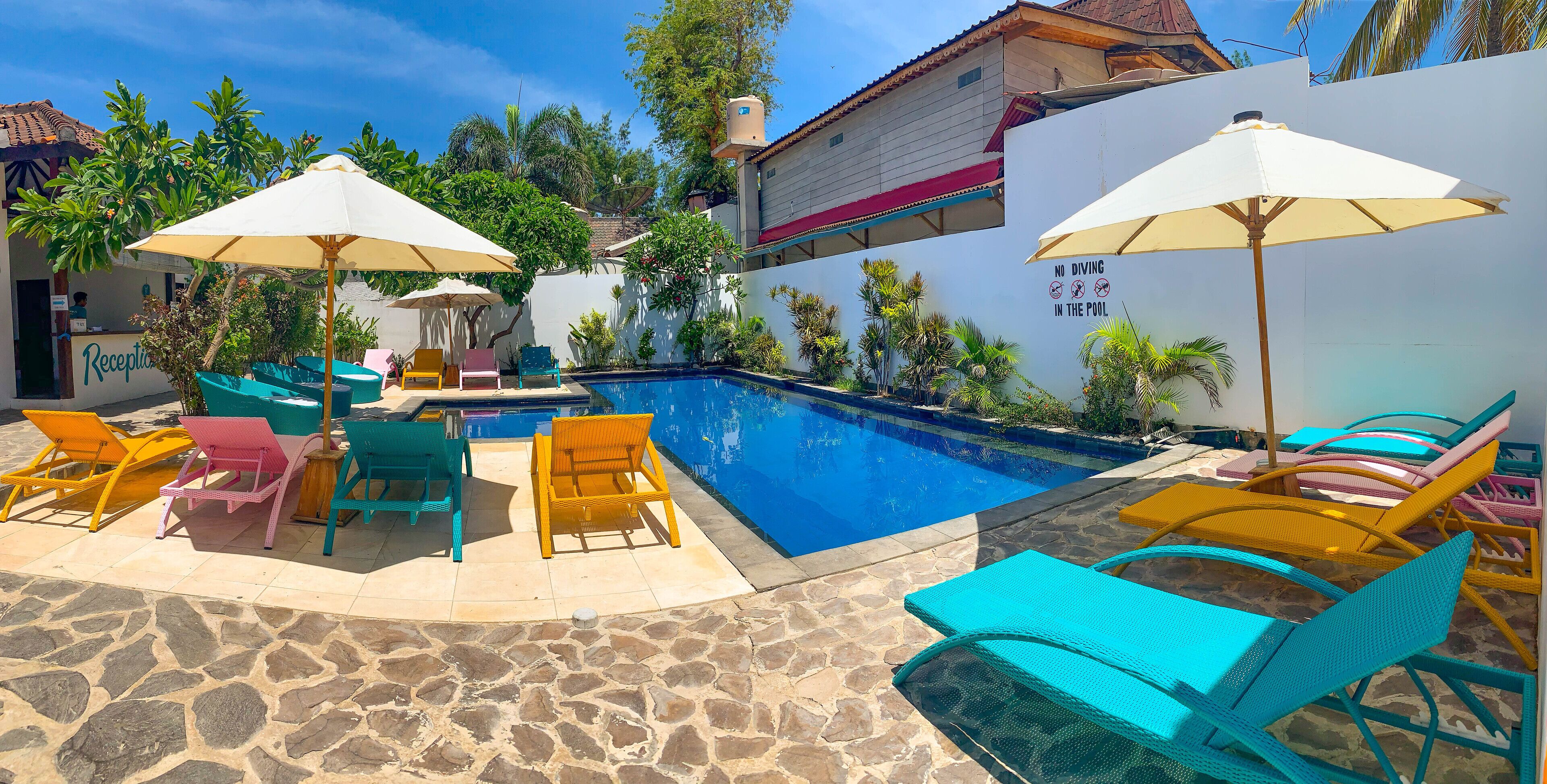 Un espace piscine avec des chaises longues et des parasols, une terrasse en pierre et un panneau indiquant « Il est interdit de plonger » sur le mur.