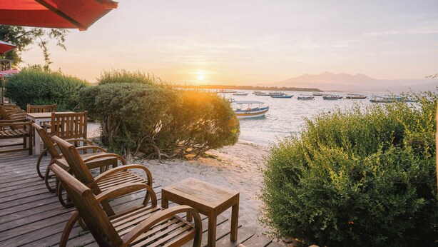 Una terraza de madera con sillas y mesas da vista a una masa de agua con barcos, una montaña al fondo y un atardecer.