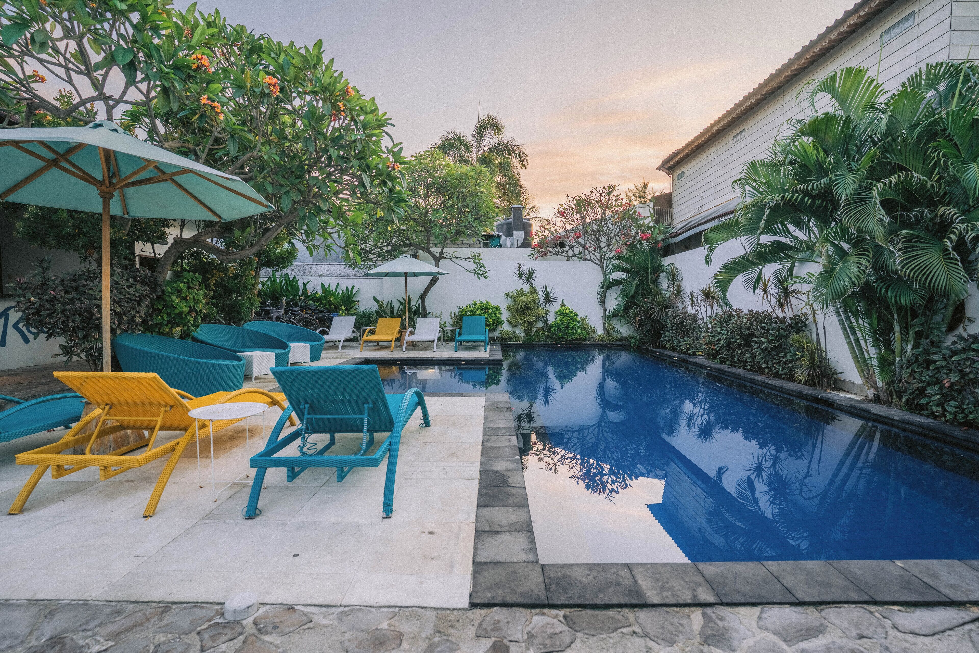 Un espace piscine doté de chaises longues et d’une petite table, entouré d’une végétation luxuriante et d’un bâtiment blanc.