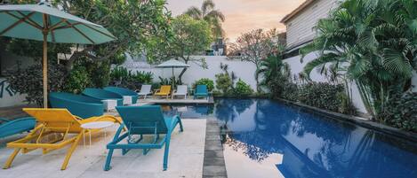 Un espace piscine doté de chaises longues et d’une petite table, entouré d’une végétation luxuriante et d’un bâtiment blanc.