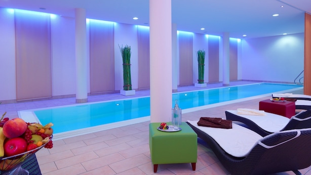 A modern indoor swimming pool area with lounge chairs, a small table with fruit, and decorative plants. - Hotel Restaurant Wessinger (Neu-Isenburg)