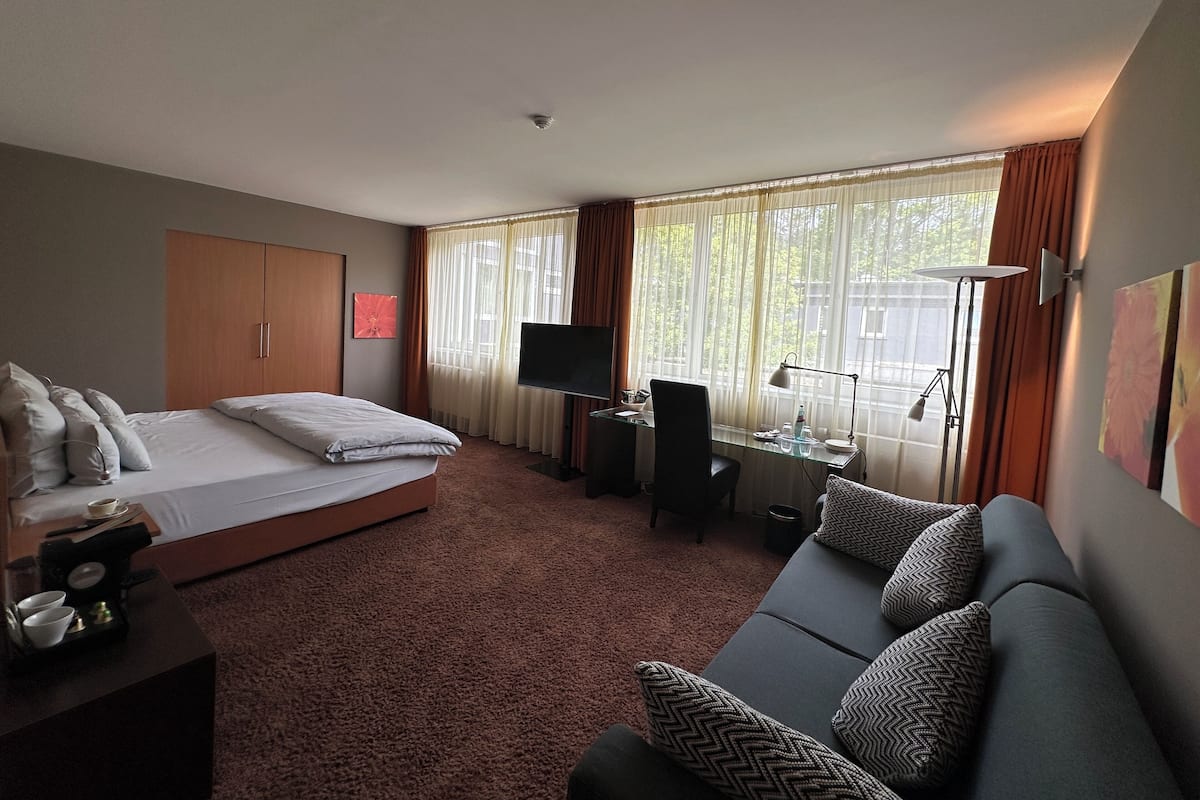 Una habitación de hotel con cama, sofá, escritorio con computadora, televisor y ventana con cortinas.