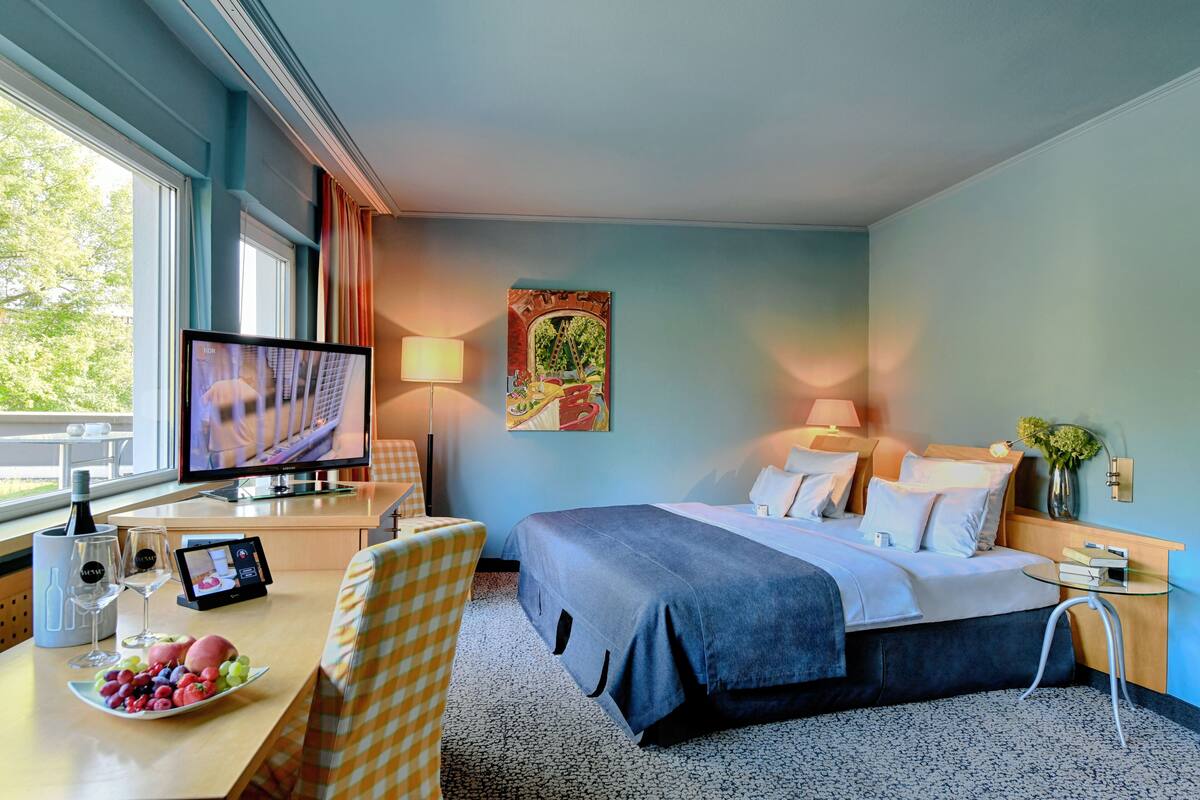 Habitación de hotel con una cama, un escritorio con computadora, televisión y una mesa de comedor con fruta.
