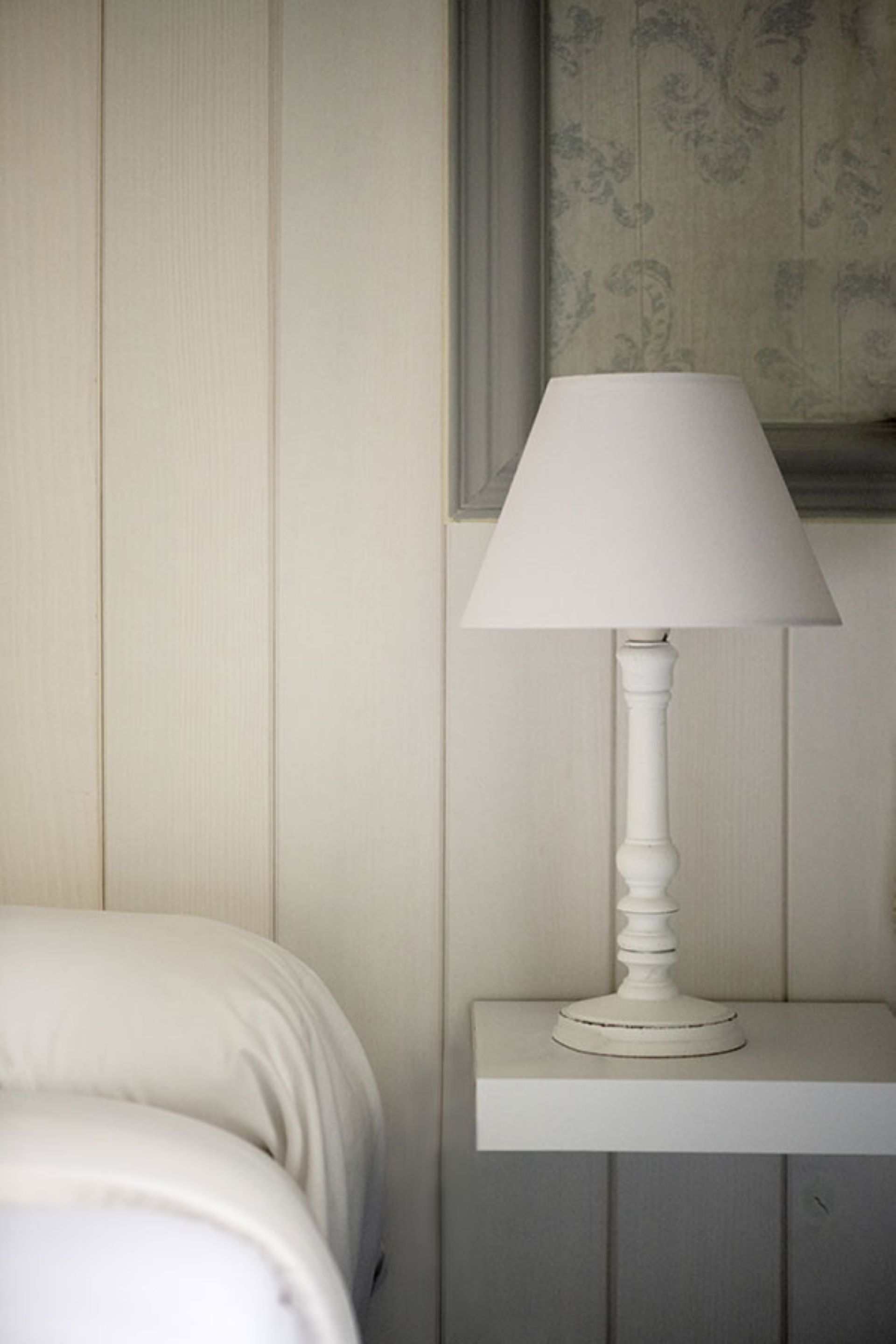 A white bedside table with a white lamp, a bed with white bedding, and a wall with a patterned wallpaper.
