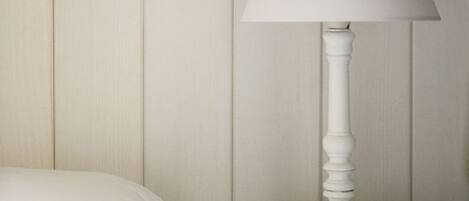 A white bedside table with a white lamp, a bed with white bedding, and a wall with a patterned wallpaper.