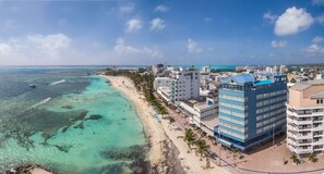 En kuststad med en strand, kristallklart turkosfärgat vatten och en framträdande blå hotellbyggnad.