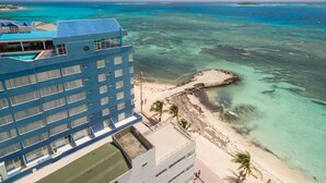 Ein Hotel mit blauer Fassade, das auf einen Strand mit klarem türkisblauem Wasser und einem Felsen überblickt.
