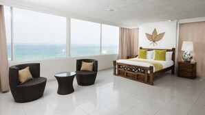 Ein modernes Hotelzimmer mit großem Fenster und Meerblick, einem hölzernen Bett mit grün-weißer Bettwäsche, zwei dunklen Sesseln und einem kleinen Tisch.
