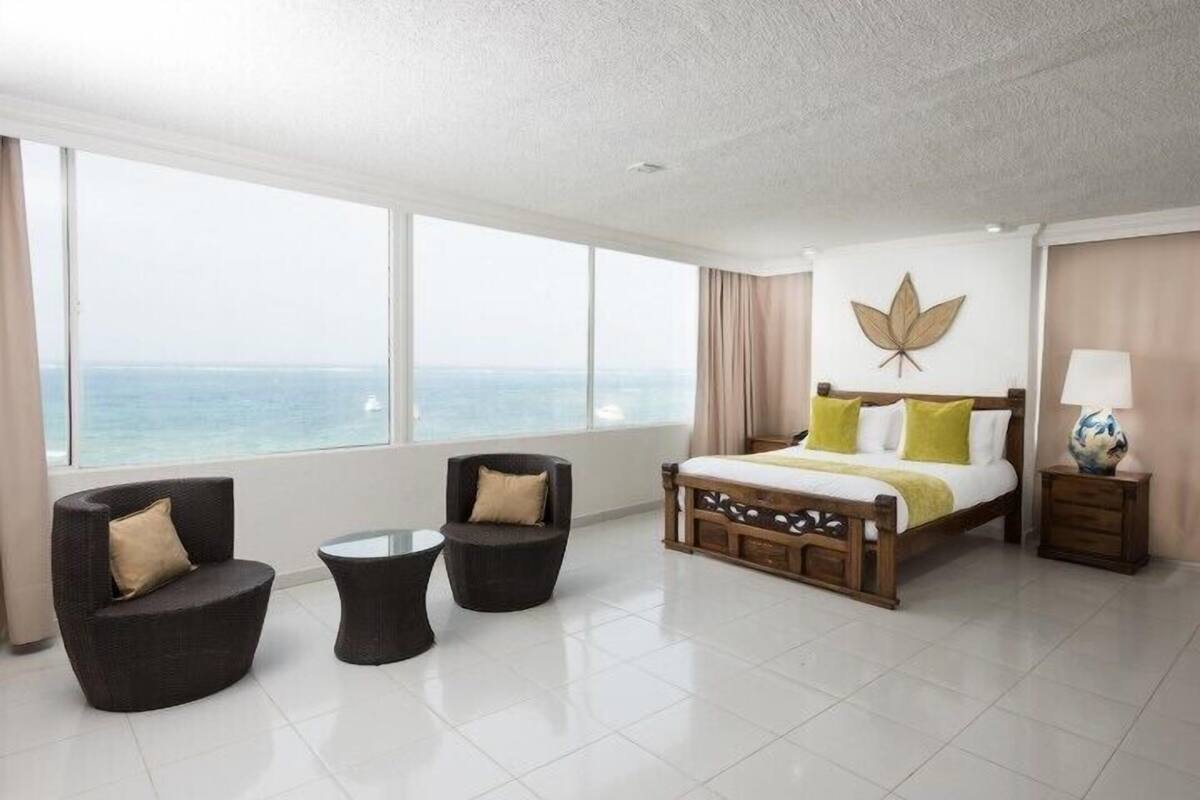 A modern hotel room with a large window offering a view of the sea, a wooden bed with green and white bedding, two dark armchairs, and a small table.