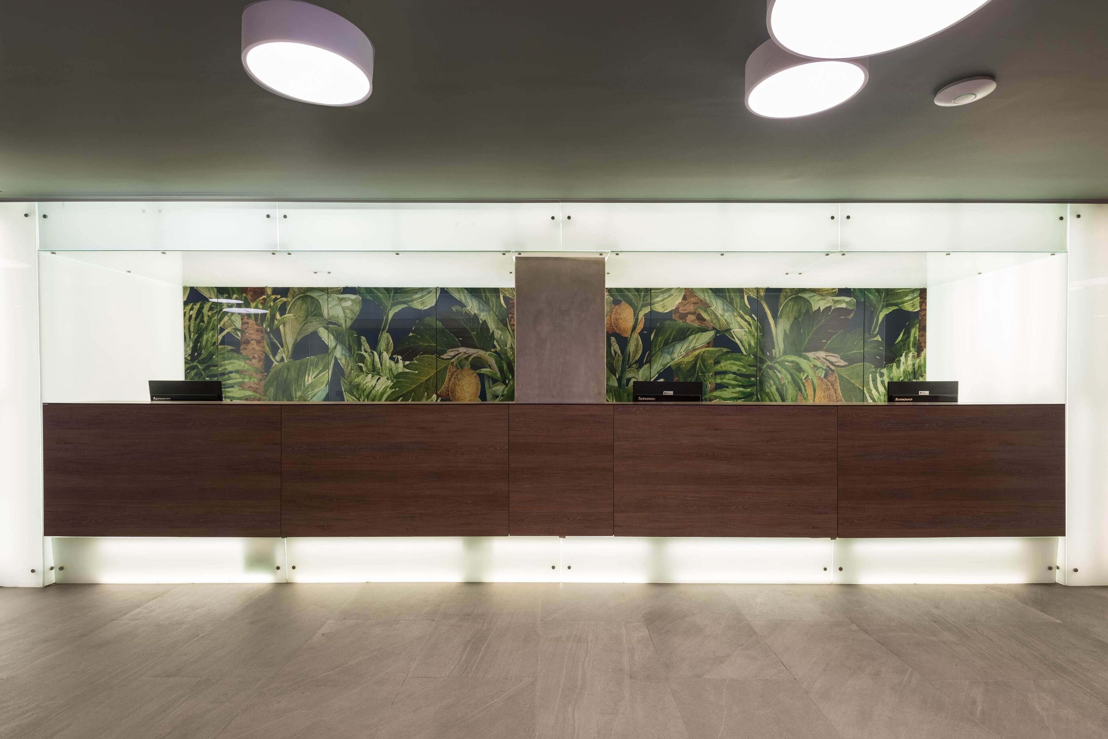 Un espace d’accueil au design moderne, comprenant un comptoir d’accueil en bois, une grande fresque murale sur le thème des régions tropicales et un éclairage encastré au plafond.
