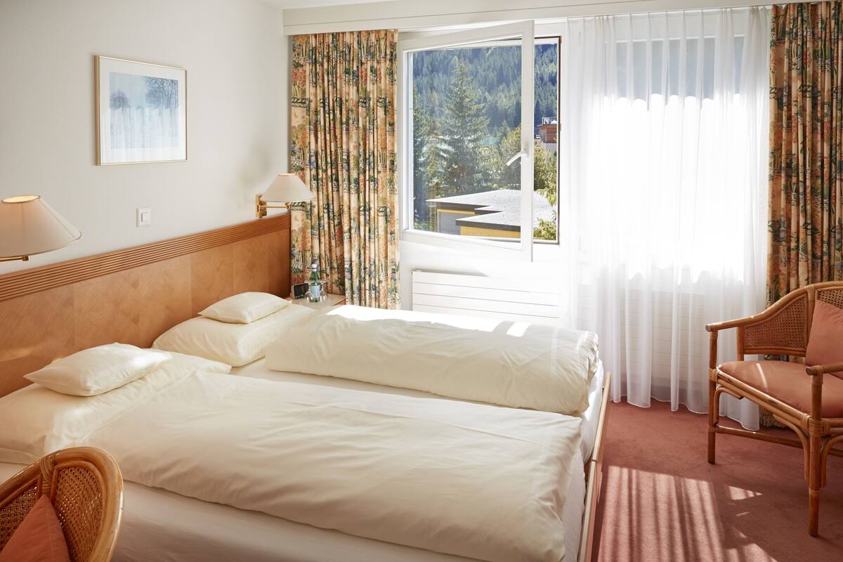 Hotellihuone, jossa on sänky, tuoli, lamppu ja ikkuna, jossa on verhot.