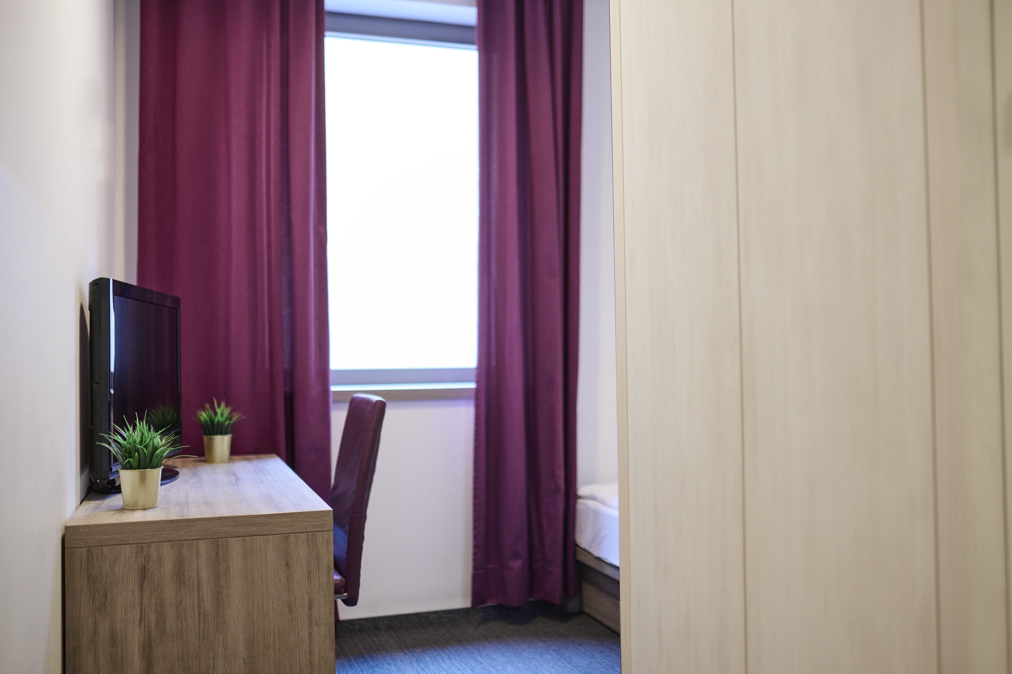 Een kleine kamer met een bureau, een stoel, een bed en een raam met paarse gordijnen.