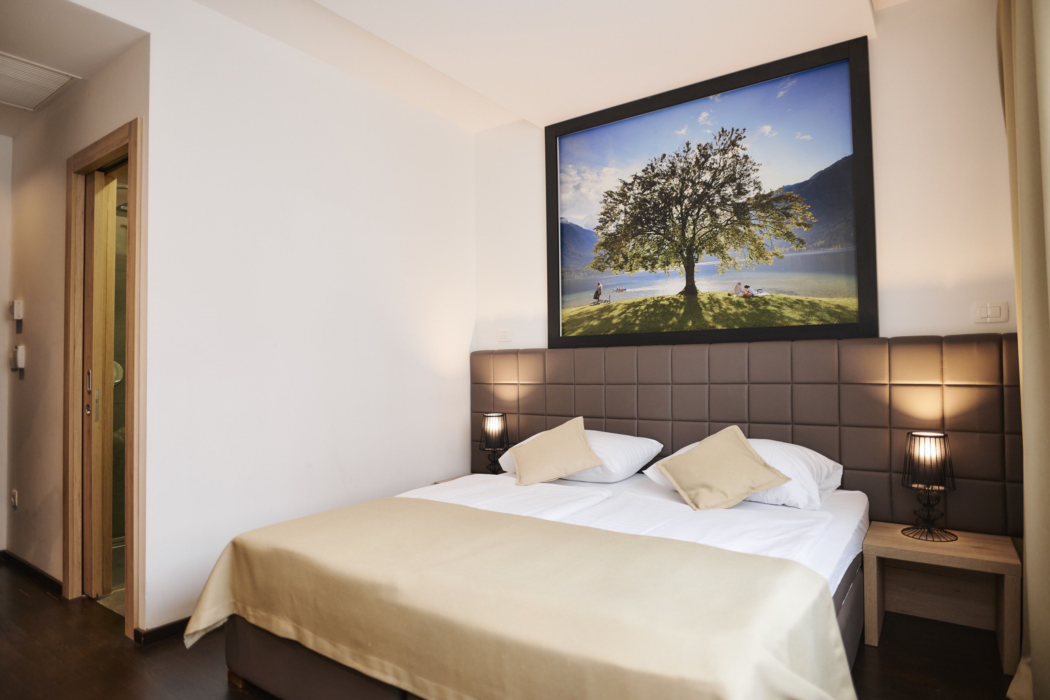 A hotel room with a large bed, two bedside tables with lamps, a painting of a tree on the wall, and a door to the left.