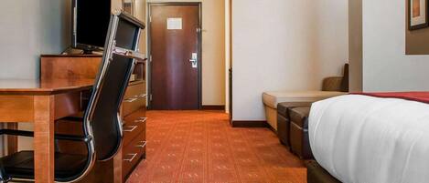 A hotel room with a bed, a desk with a computer and chair, a small sofa, and a door.