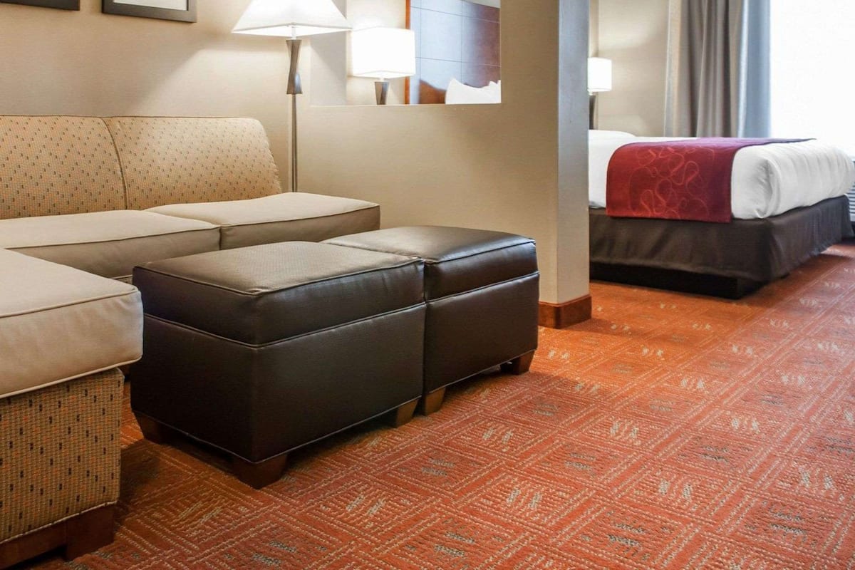 Ein Hotelzimmer mit einer Couch, einem Hocker und einem Bett mit roter Decke. An der Wand hängen zwei gerahmte Bilder und auf dem Boden steht eine Lampe.