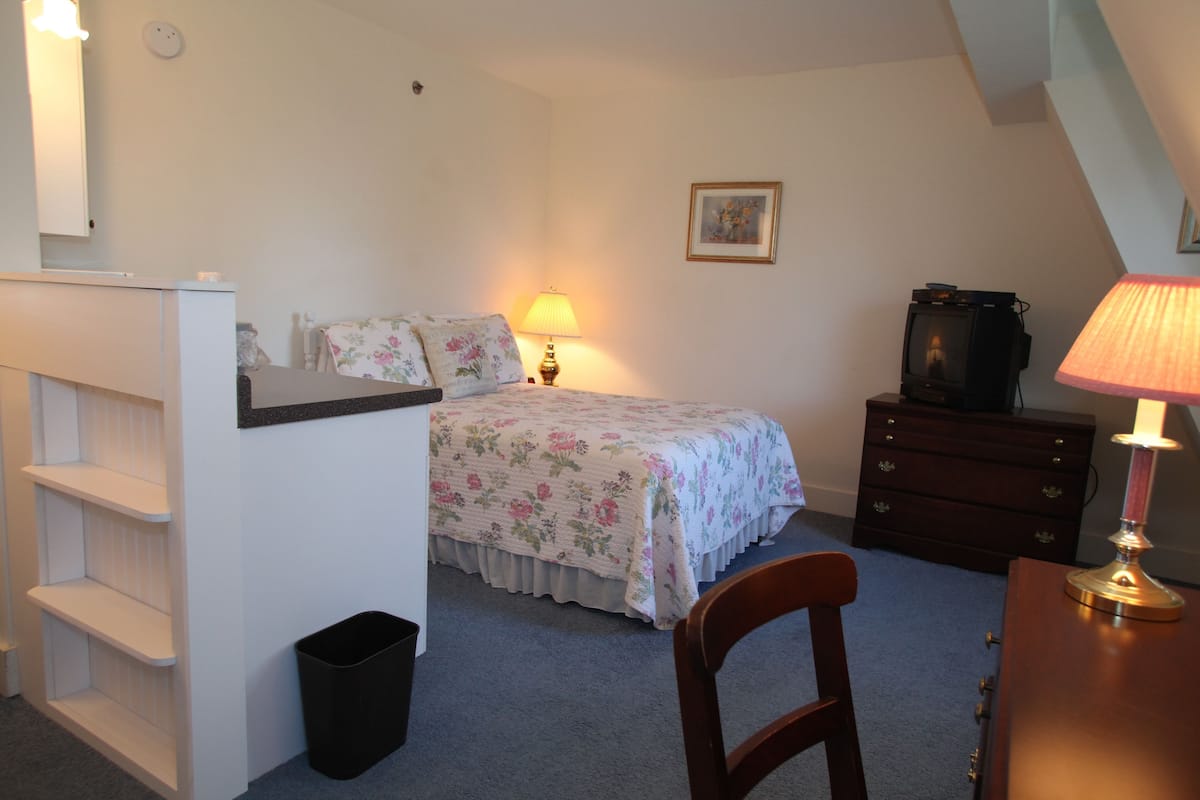 A bedroom with a bed, a desk, a chair, a dresser, a lamp, a trash bin, and a picture on the wall.