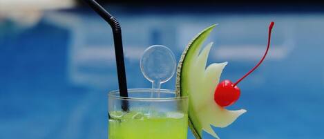 A tall glass with a lime-colored drink, a black straw, and a cherry garnish on a white table by a pool.