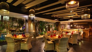 A restaurant with wicker furniture and hanging lights, featuring a bar area and indoor plants.