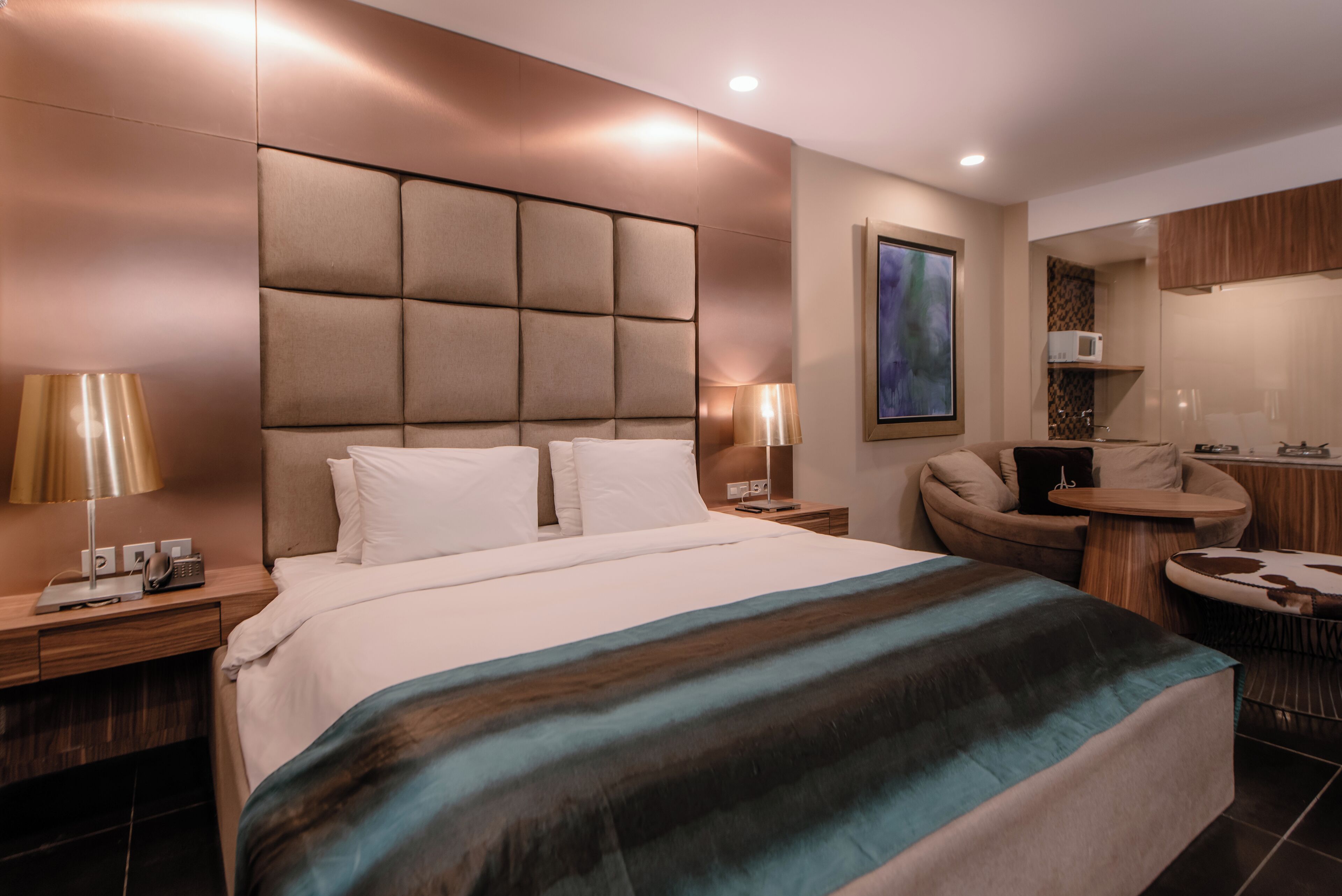 A modern hotel room with a large bed, a desk, a chair, and a small kitchenette.