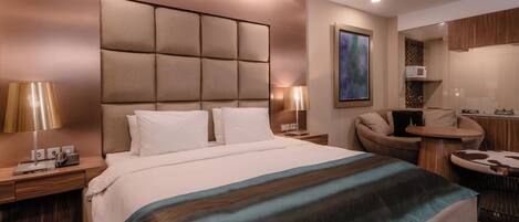 A modern hotel room with a large bed, a desk, a chair, and a small kitchenette.