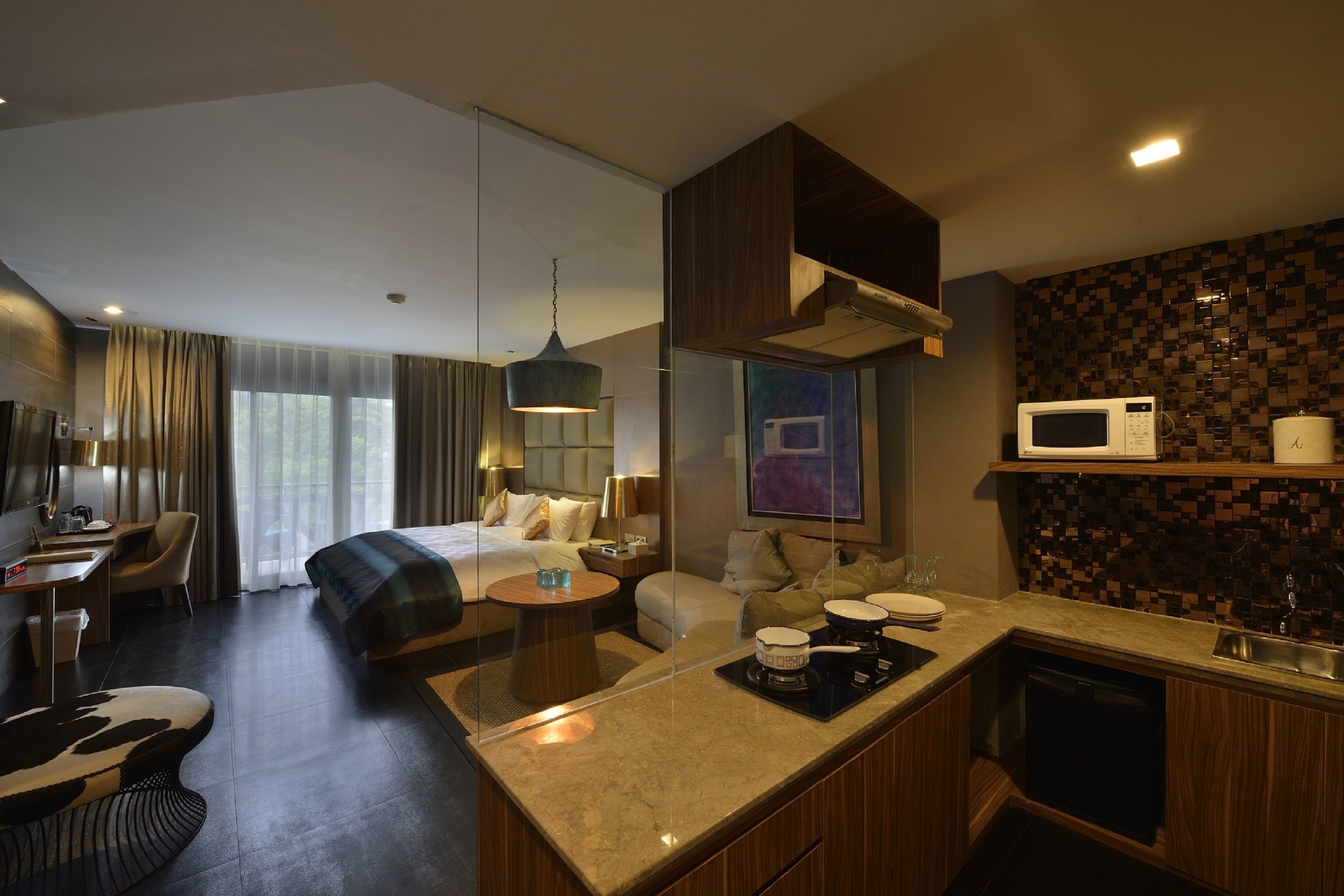 A modern hotel room with a bed, a desk, a sofa, a dining area, and a kitchen with a microwave.