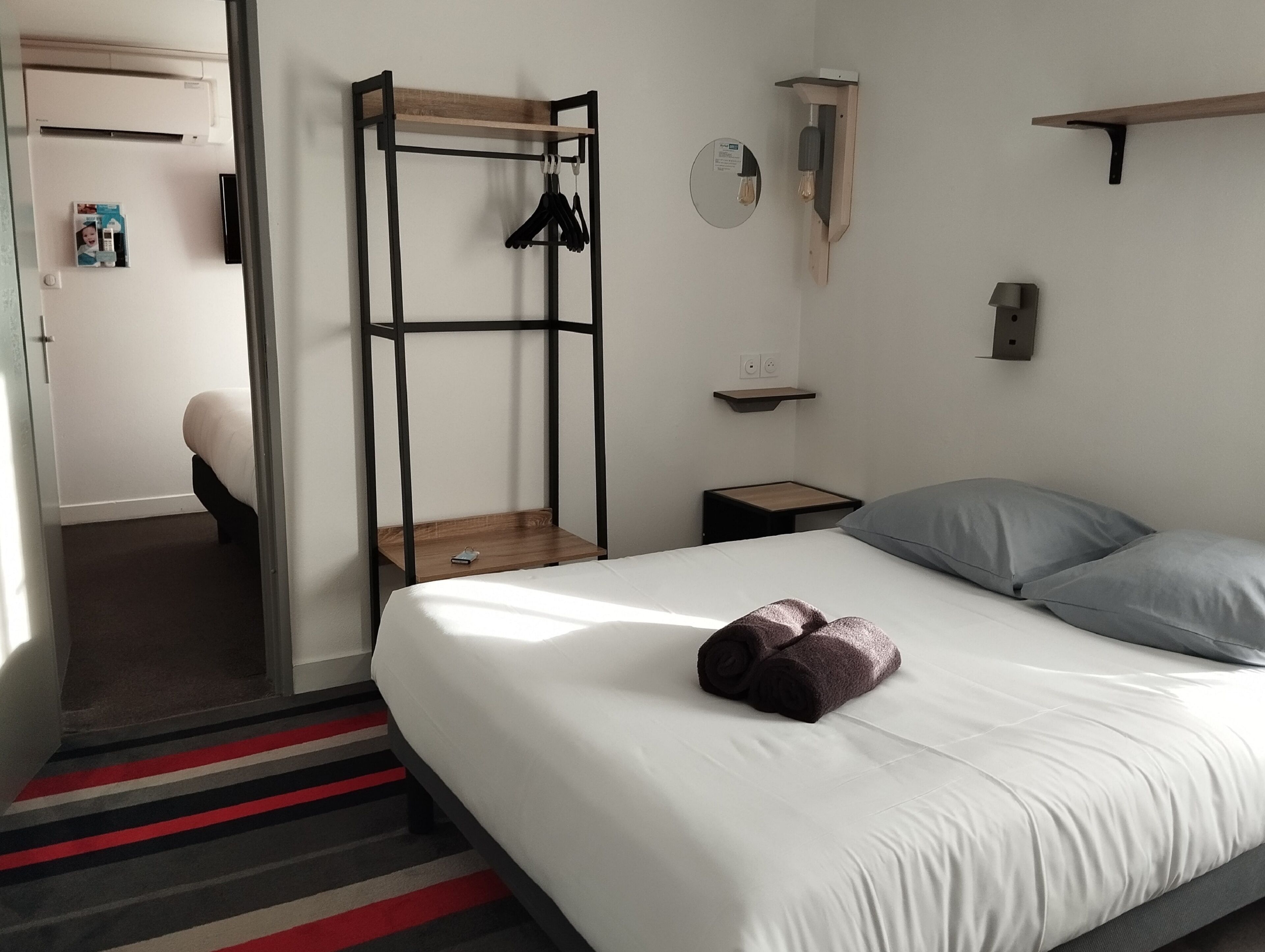 A bedroom with a bed, a wooden bench, a wall-mounted light fixture, and a small shelf.