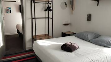 A bedroom with a bed, a wooden bench, a wall-mounted light fixture, and a small shelf.