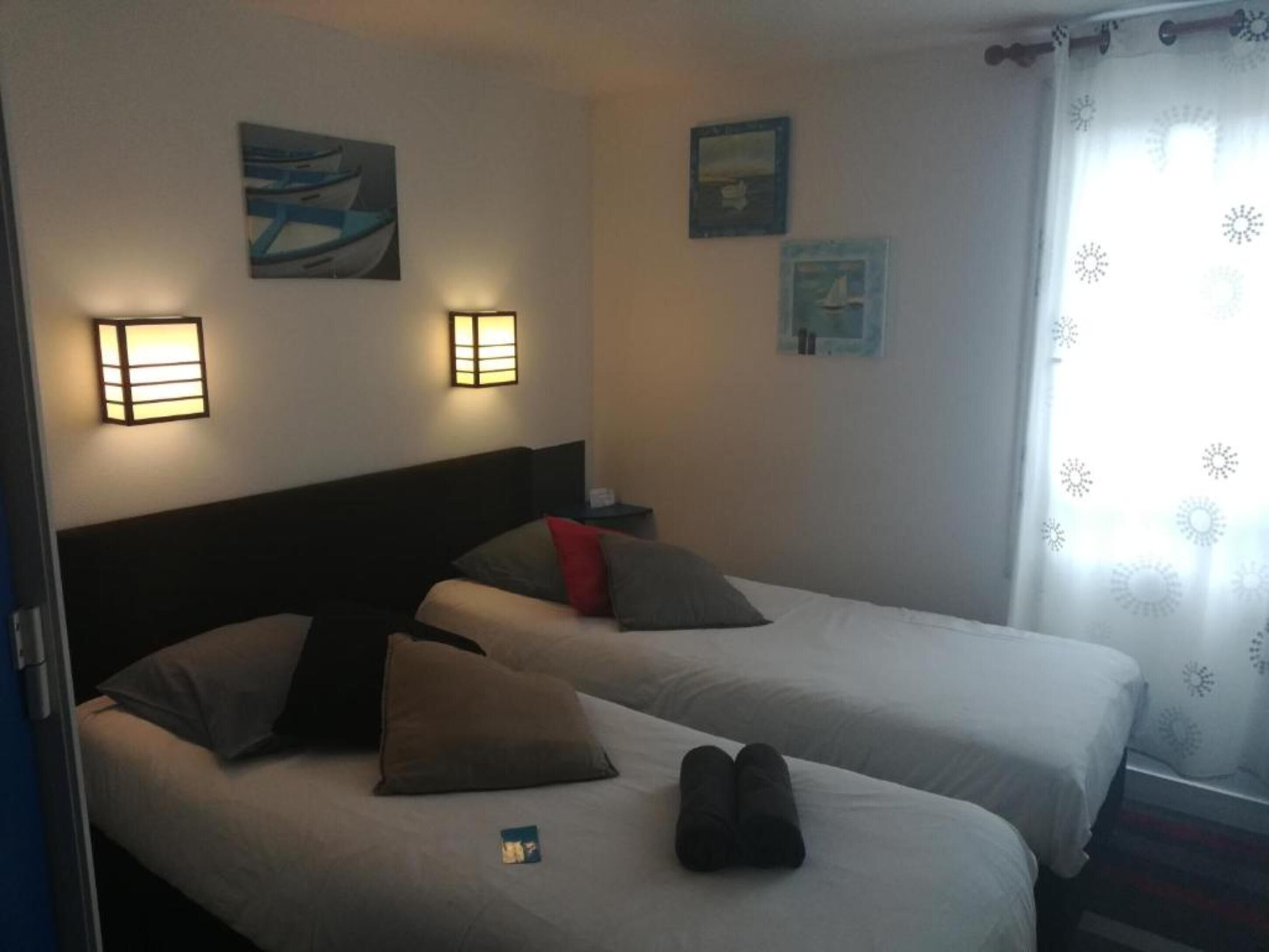 Habitación de hotel con dos camas, cada una con almohadas y una manta. Hay luces fijadas a la pared y cuadros enmarcados.