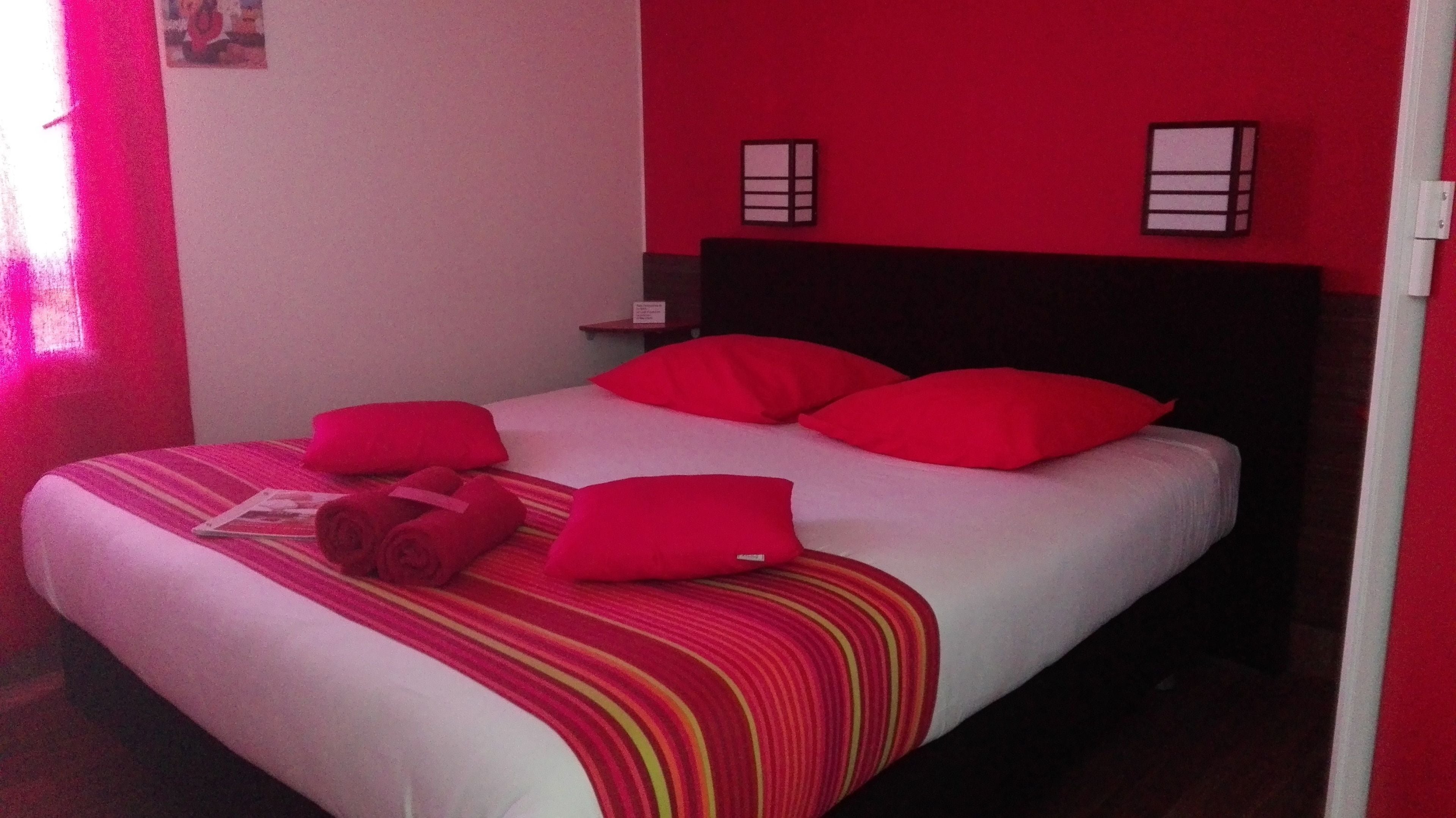 Un lit avec des oreillers rouges et une couverture à rayures, une table de chevet avec une lampe, et un mur rouge orné de deux appliques murales.