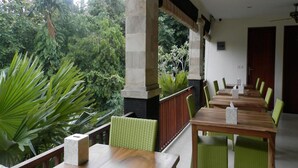 Un espacio de comedor al aire libre con mesas de madera y sillas verdes, rodeado de abundante vegetación y un pilar de piedra.