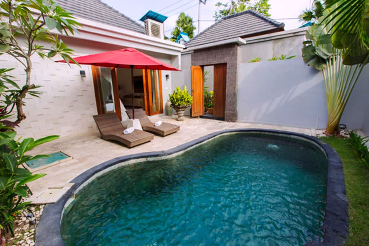 Ein Swimmingpool mit Liegestühlen und einem roten Sonnenschirm, umgeben von einem kleinen Garten und einem weißen Gebäude.