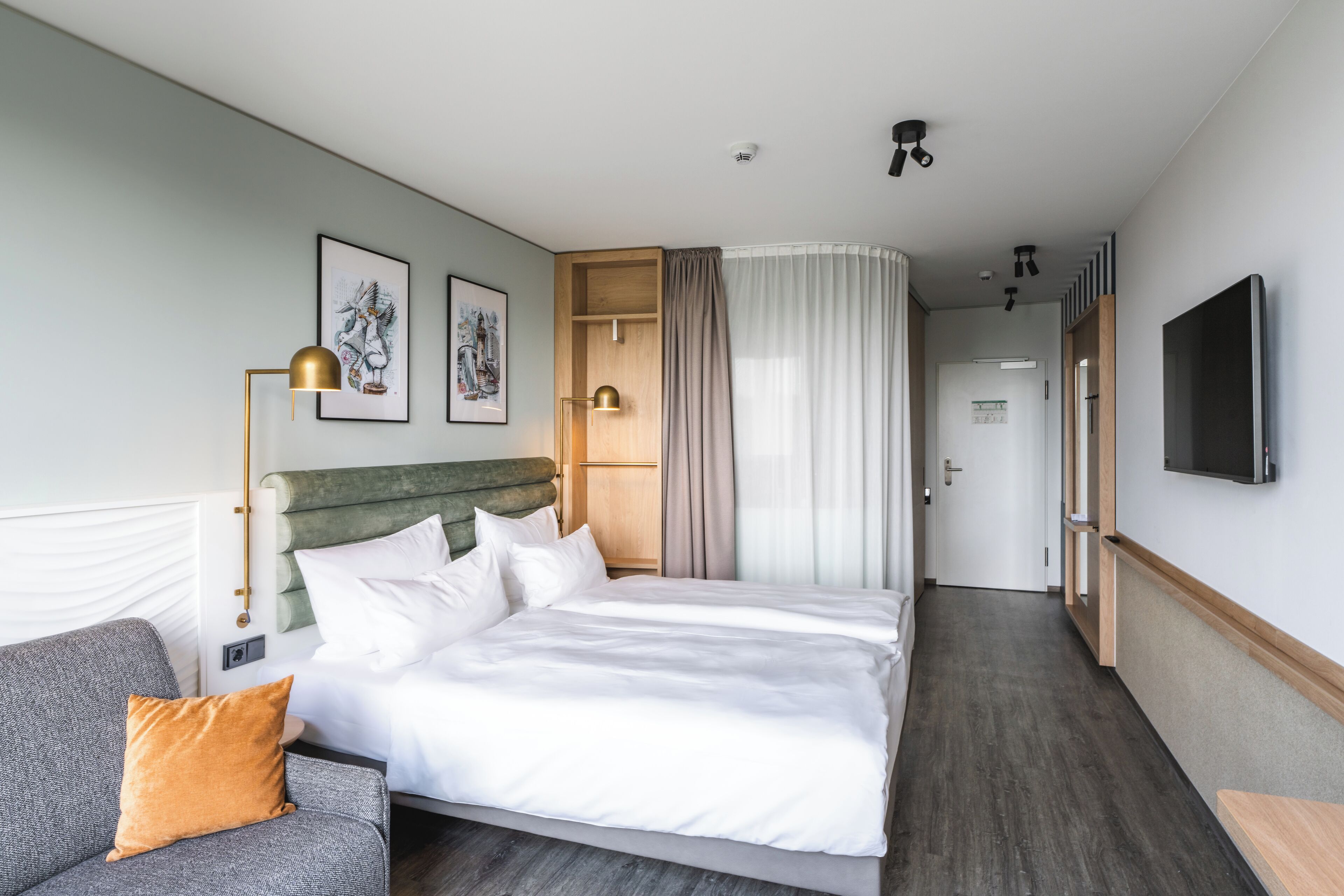 A modern hotel room with a large bed, a grey sofa, a flat-screen TV, and a wooden headboard.