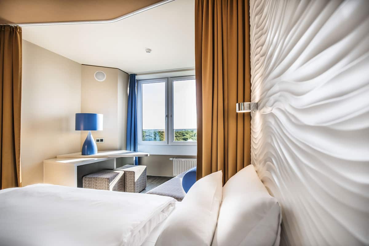 A hotel room with a bed, a desk, a chair, and a view of the outdoors through a window.