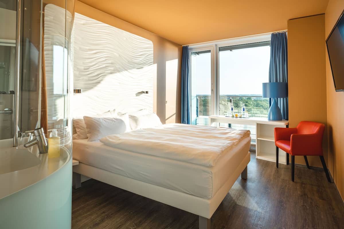 A modern hotel room with a large bed, a bathroom with a sink, and a view of the outdoors through a window.