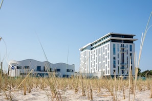 A modern hotel with a flat roof and multiple floors, surrounded by sand and tall grass.