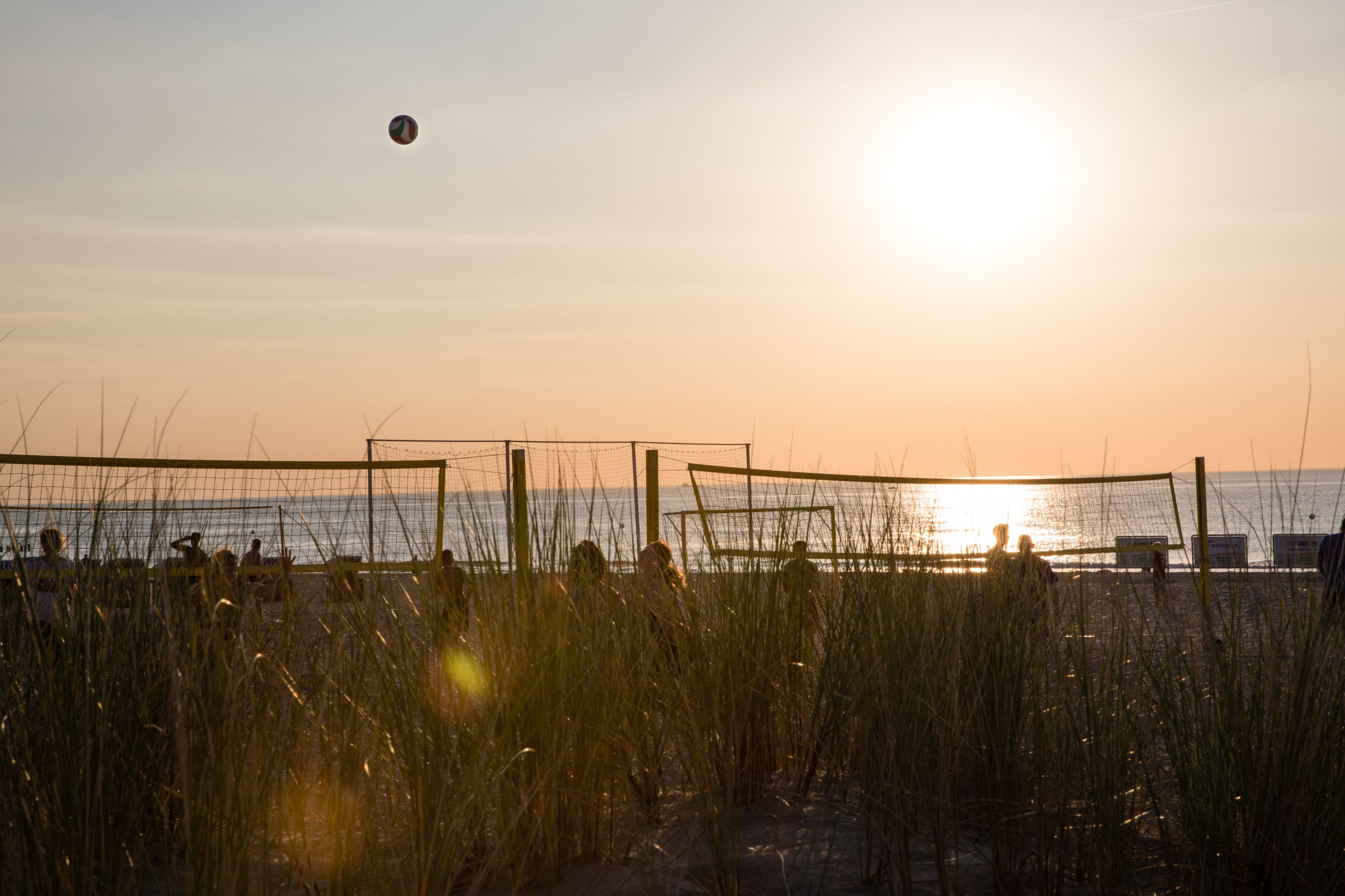 Una red de voleibol de playa con jugadores al fondo, un atardecer y un mar tranquilo.