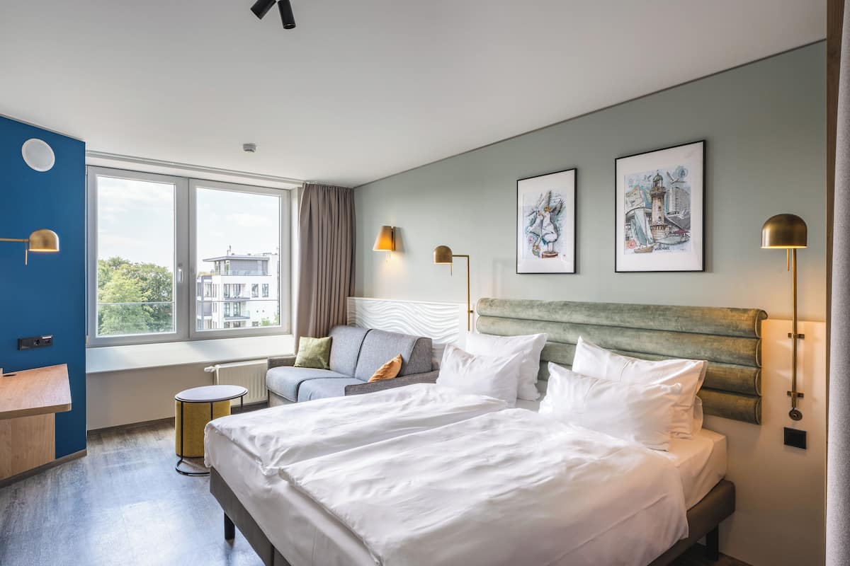 A modern hotel room with a large bed, a sofa, a small round table, and two wall-mounted lamps.