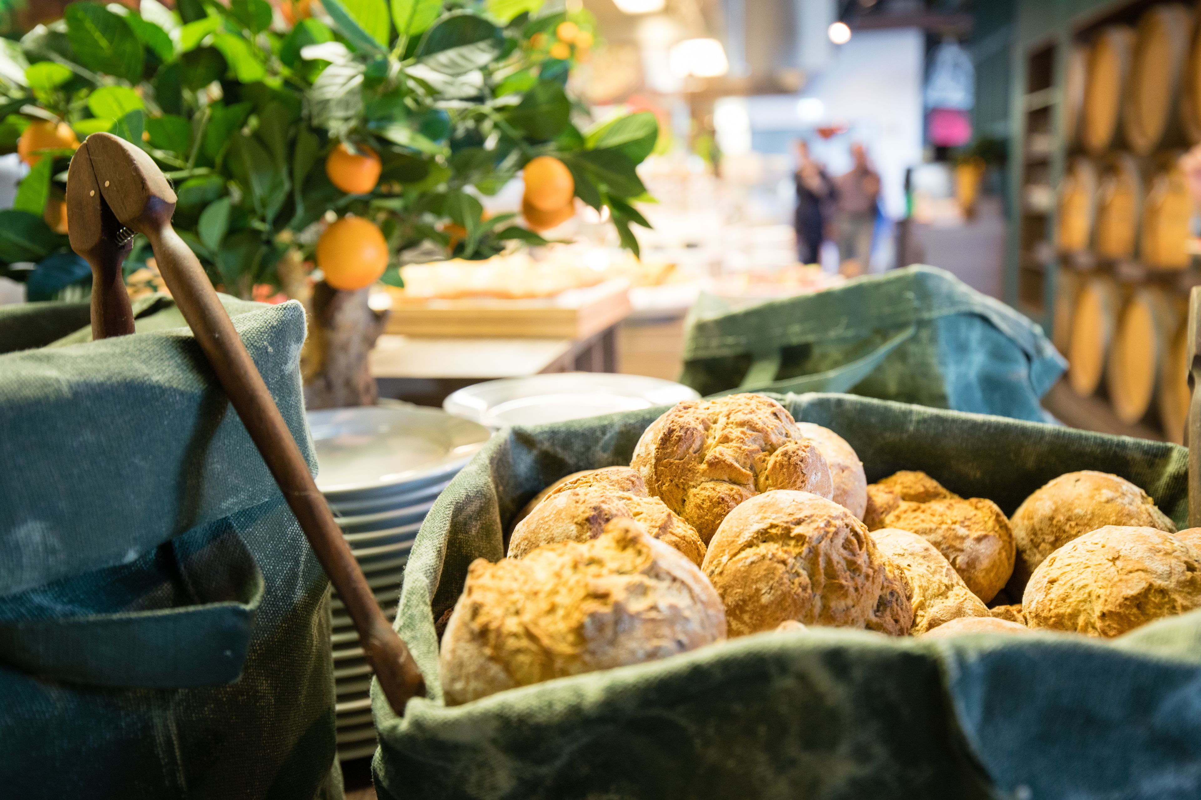 A basket of freshly baked bread, a wooden mallet, and a potted plant with oranges in a market setting.